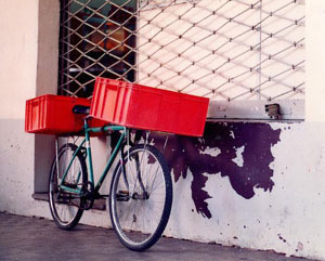 Foto de uma bicicleta de carga verde com caixas laranjas encostada numa parede
